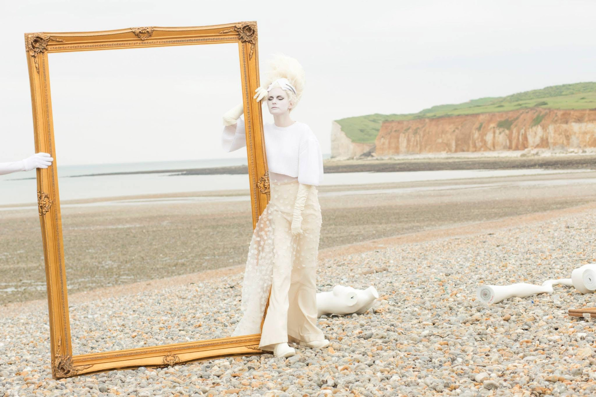 Large ornate gold frame on a pebble beach with a pale-dressed model.